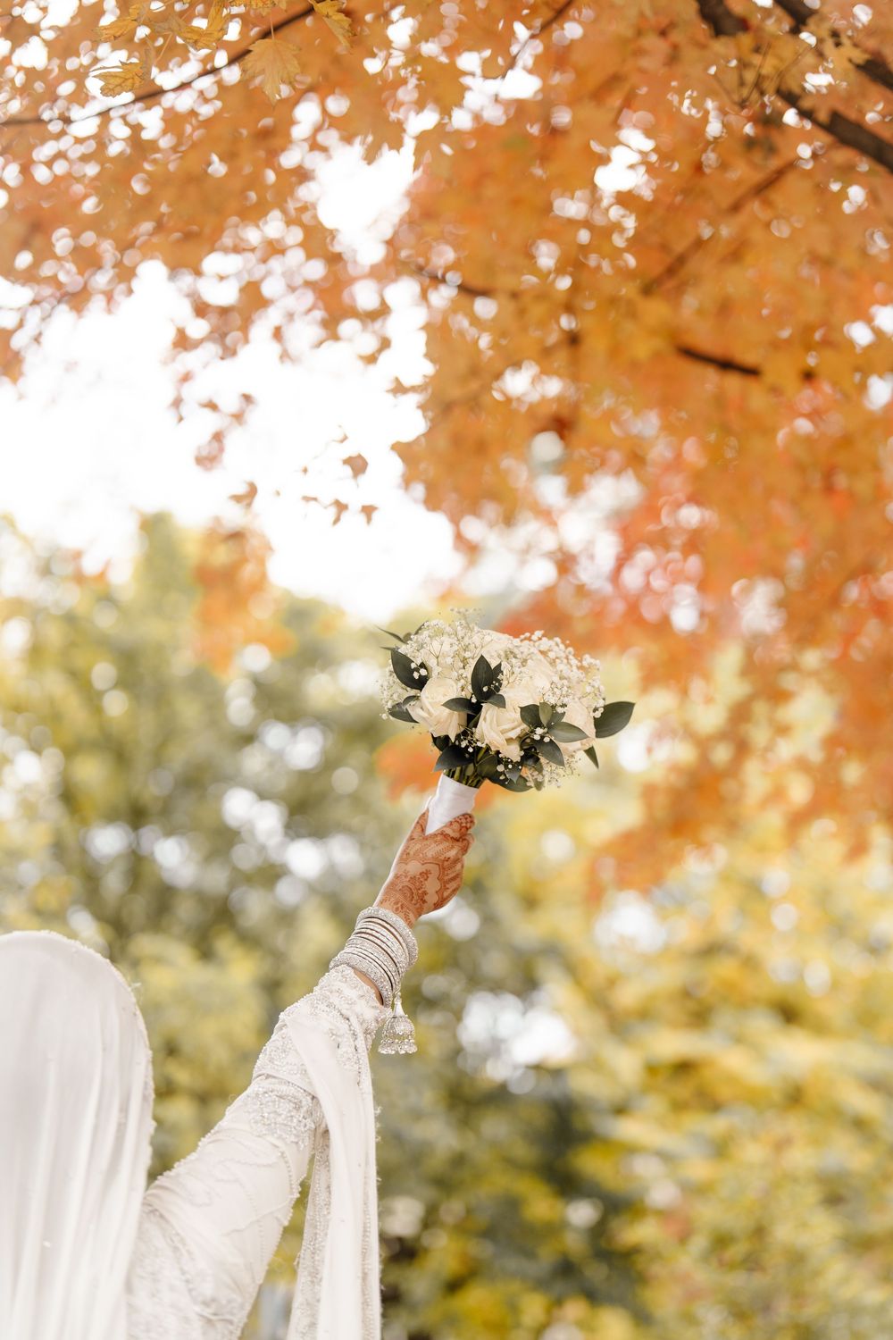 South Asian bride with henna-adorned hand raises white floral bouquet beneath vibrant orange autumn maple tree.