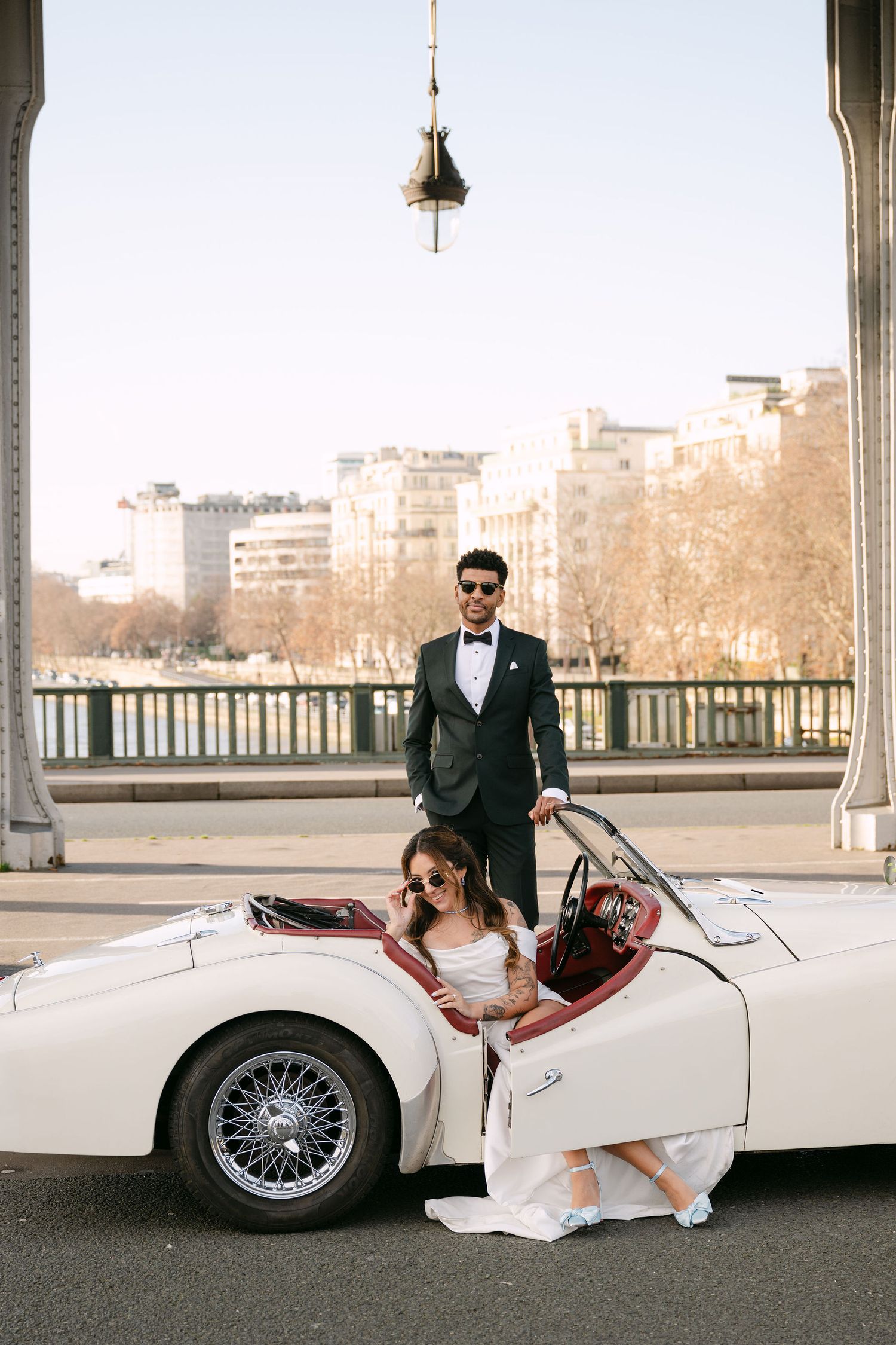Elegant wedding couple poses with vintage white convertible car under covered pavilion at sunset.