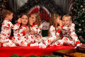 Children in matching red and white Christmas pajamas lie together on a red blanket during holiday photo session.