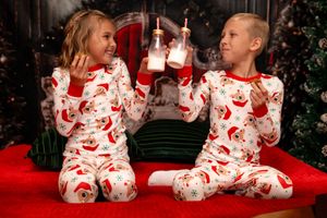 Two children in matching Santa-patterned pajamas sit on a red blanket holding Christmas drinks during holiday photoshoot.