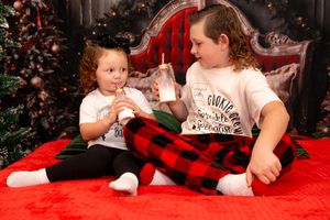 Children in matching holiday shirts sit on a red blanket in front of a decorated Christmas tree.