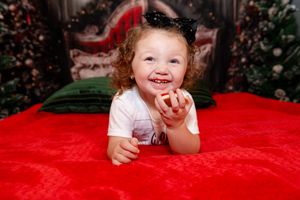 Child with curly red hair laughs while lying on bright red blanket during holiday photo session.