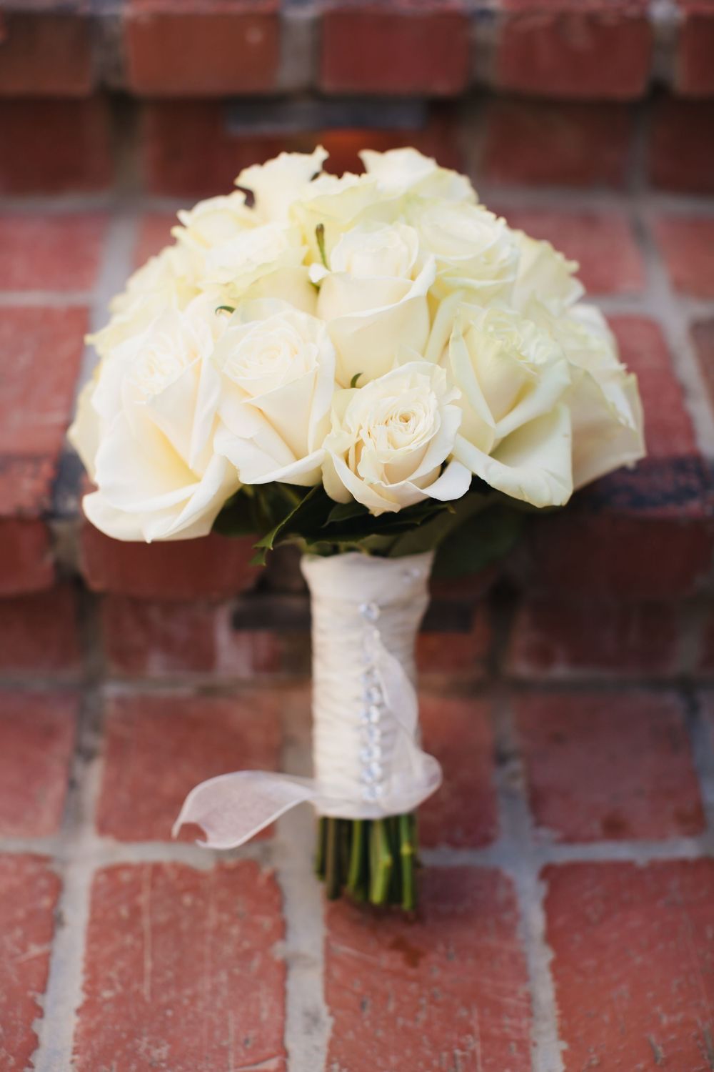 White rose wedding bouquet wrapped in lace ribbon rests on red brick surface.