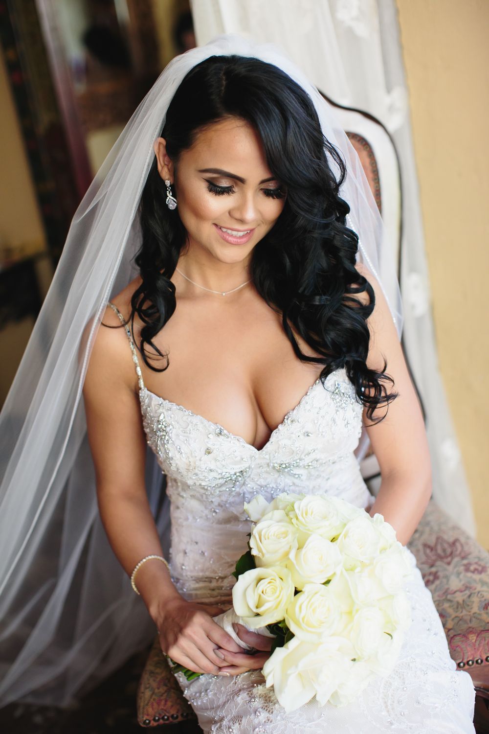 Beautiful bride in white lace wedding gown holds cream roses while wearing long veil and elegant jewelry.
