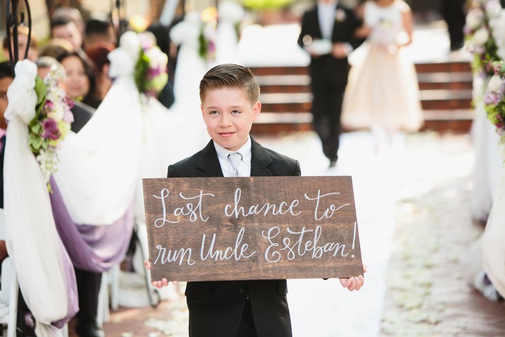 Wedding party members hold rustic wooden signs with playful messages during outdoor ceremony.