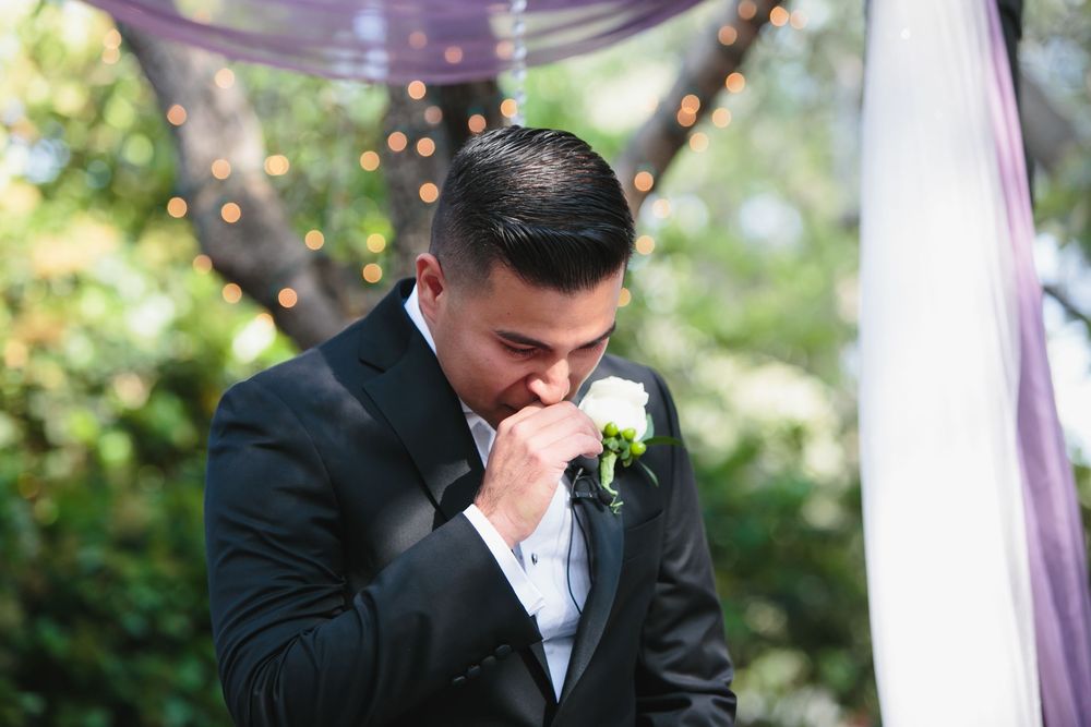 Groom wipes away tears during an emotional outdoor wedding ceremony beneath purple fabric draping.