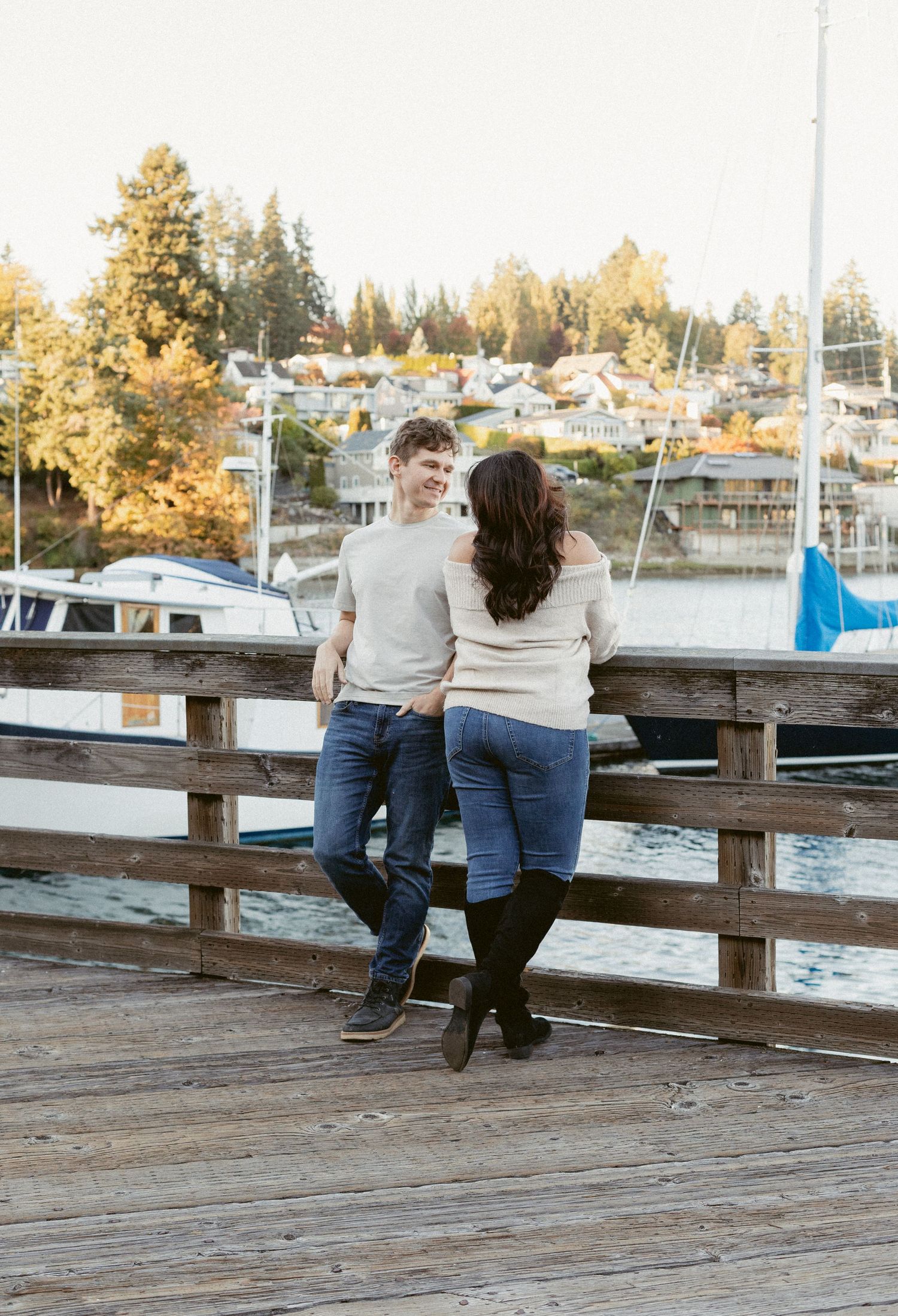 A couple sits together on a wooden dock overlooking a marina with boats and autumn trees in the background.