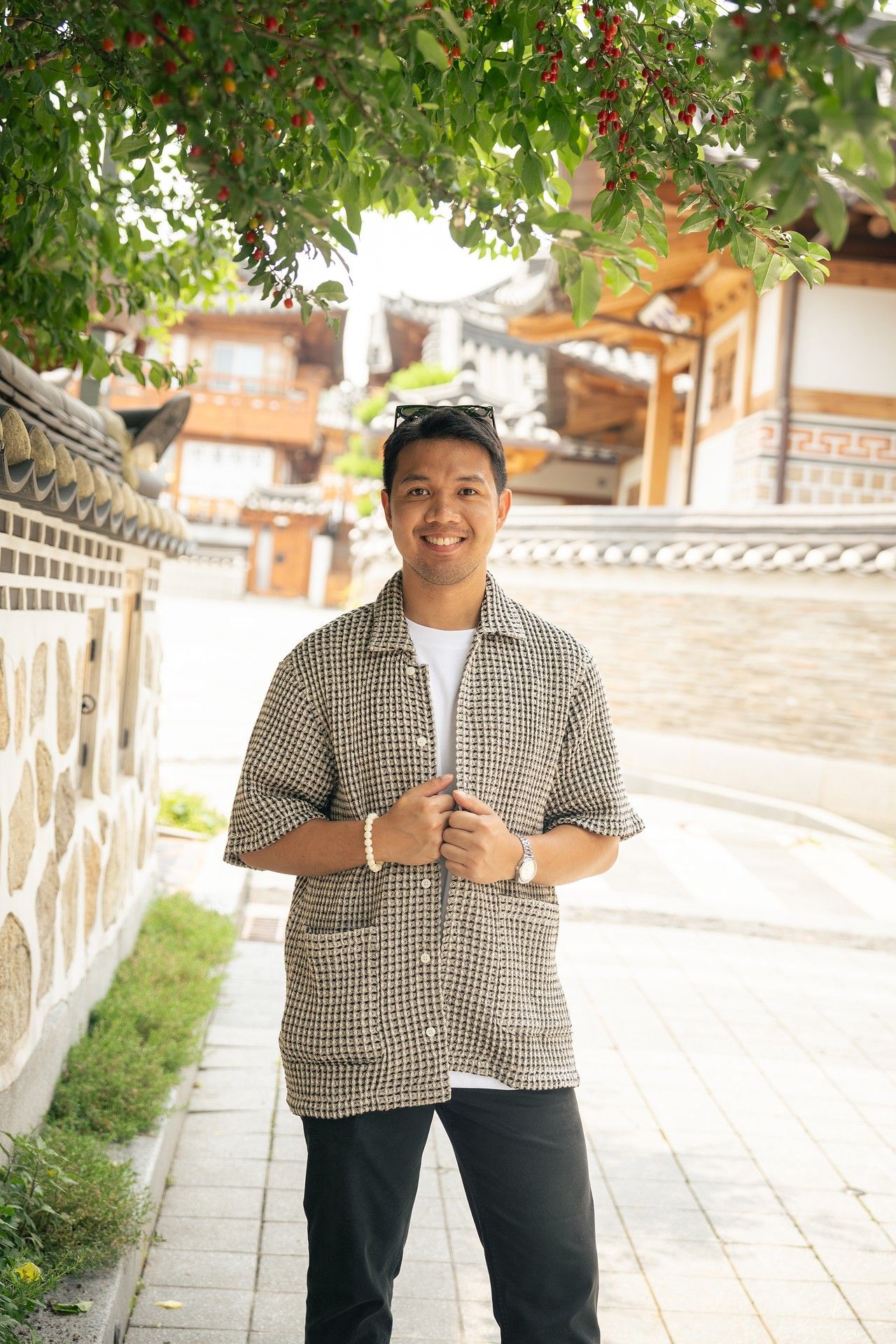 Person wearing a checkered shirt and dark pants stands on a stone path near traditional Asian architecture.