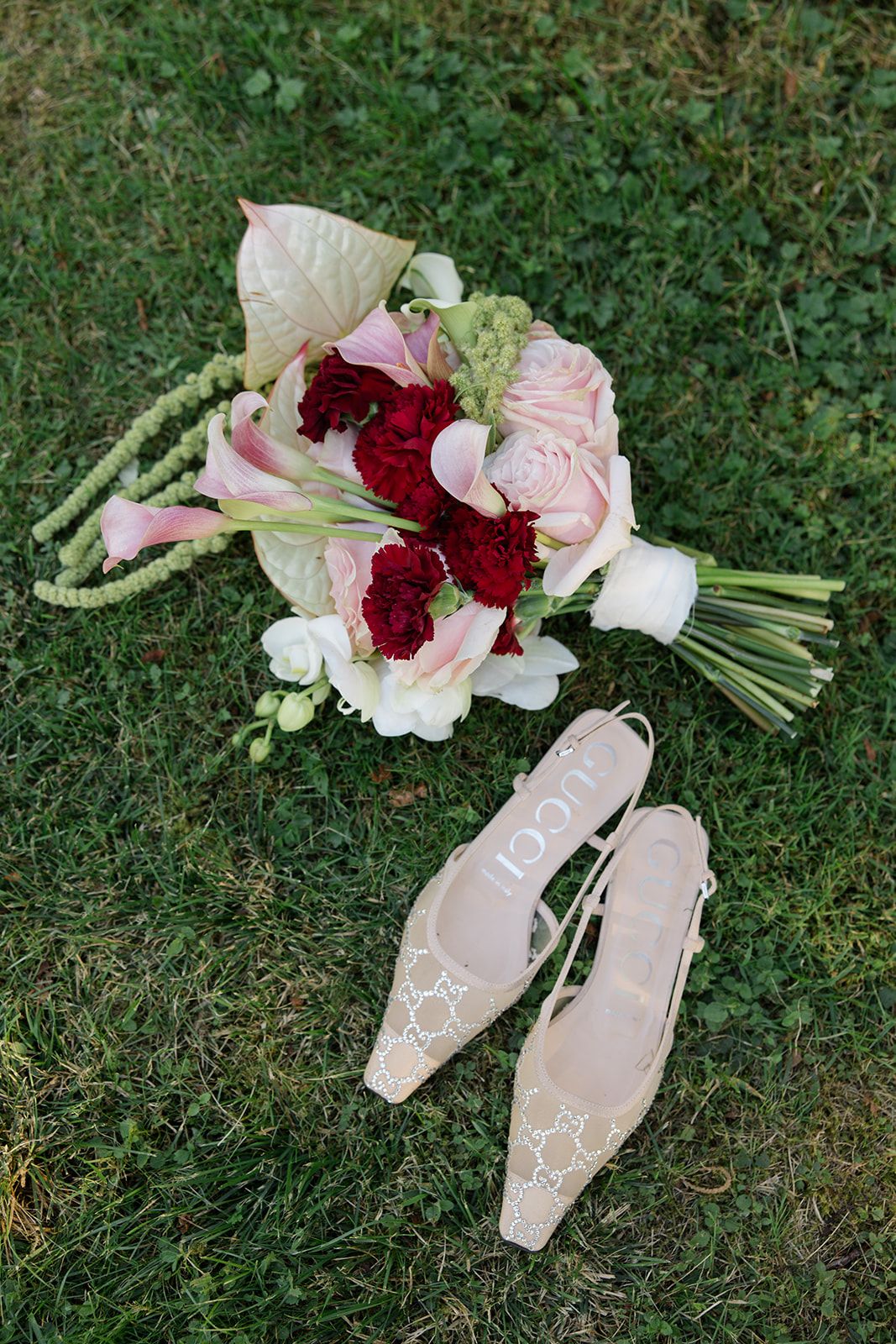 A bridal bouquet of pink and deep burgundy flowers rests on green grass next to delicate blush-colored shoes.