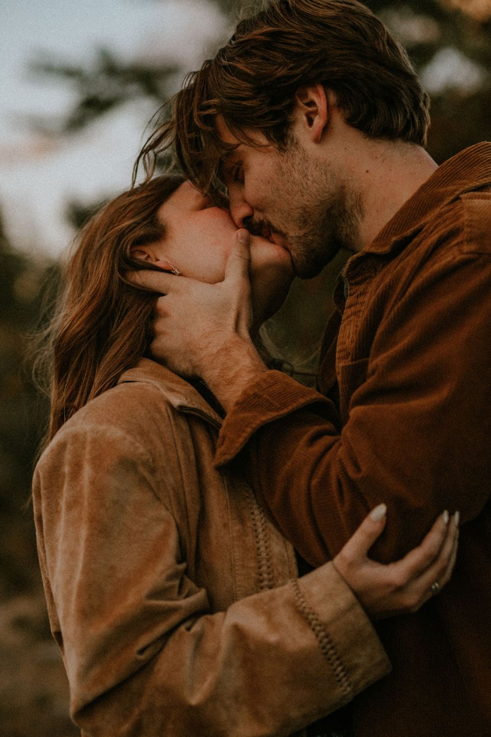 A romantic couple in brown jackets share an intimate moment at sunset in moody autumn lighting.