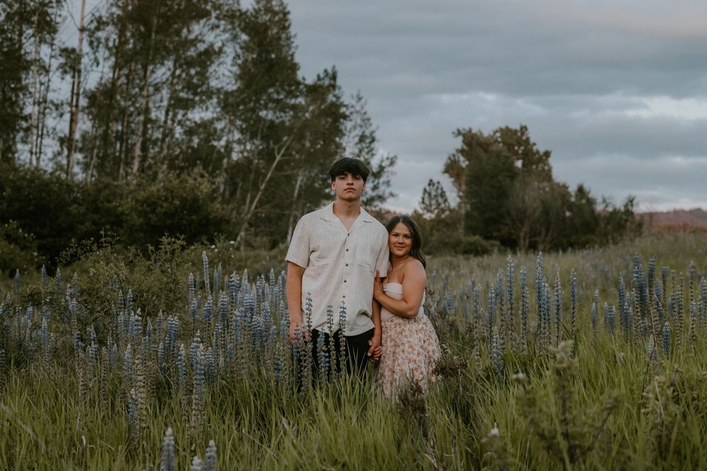 A couple shares a tender moment together in a field of tall grass at sunset with moody clouds overhead.