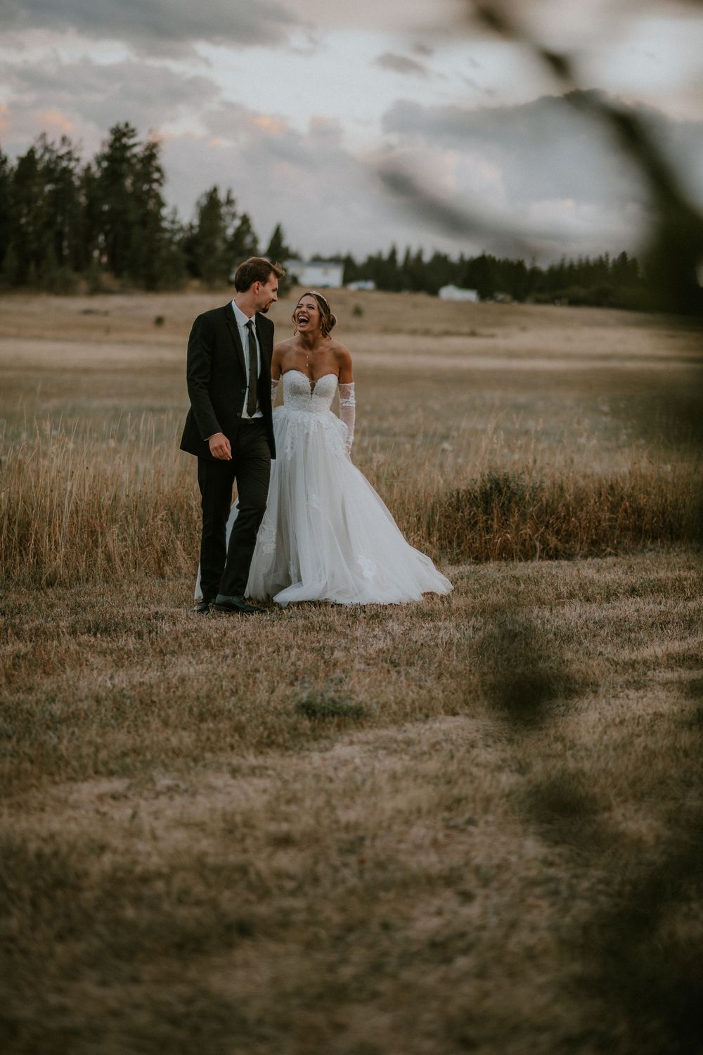A couple in wedding attire stands together in a rustic field at dusk with evergreen trees in the background.