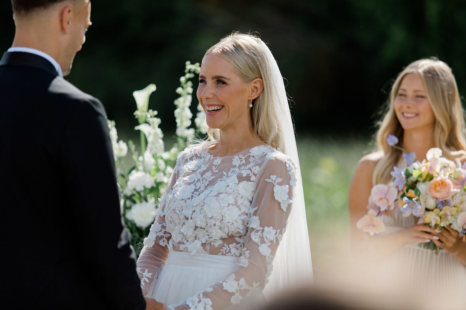 A bride and bridesmaids in floral dresses laugh together during an outdoor wedding photo session.