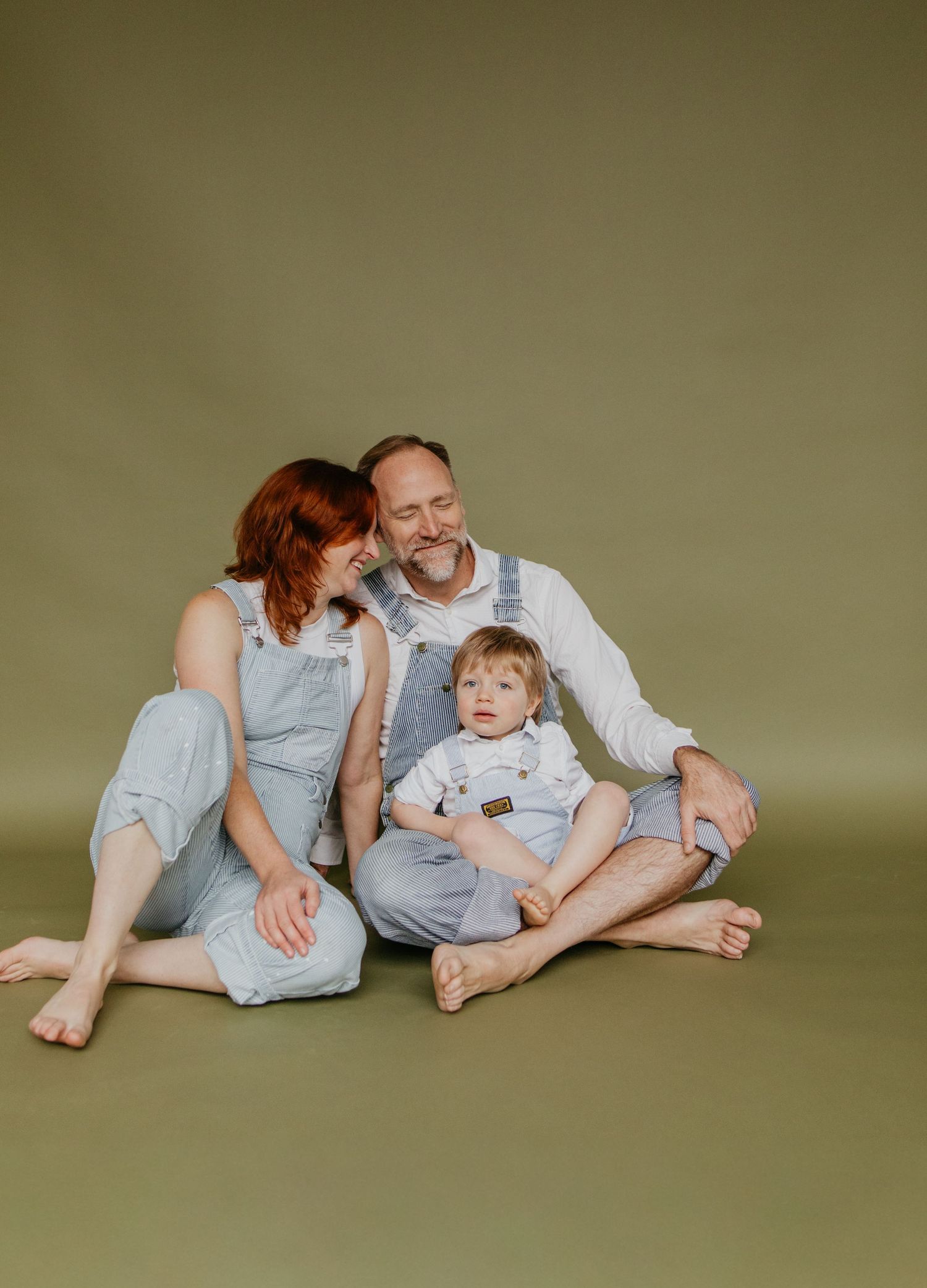 A family sits together on the floor wearing coordinated blue and white outfits against an olive green backdrop.