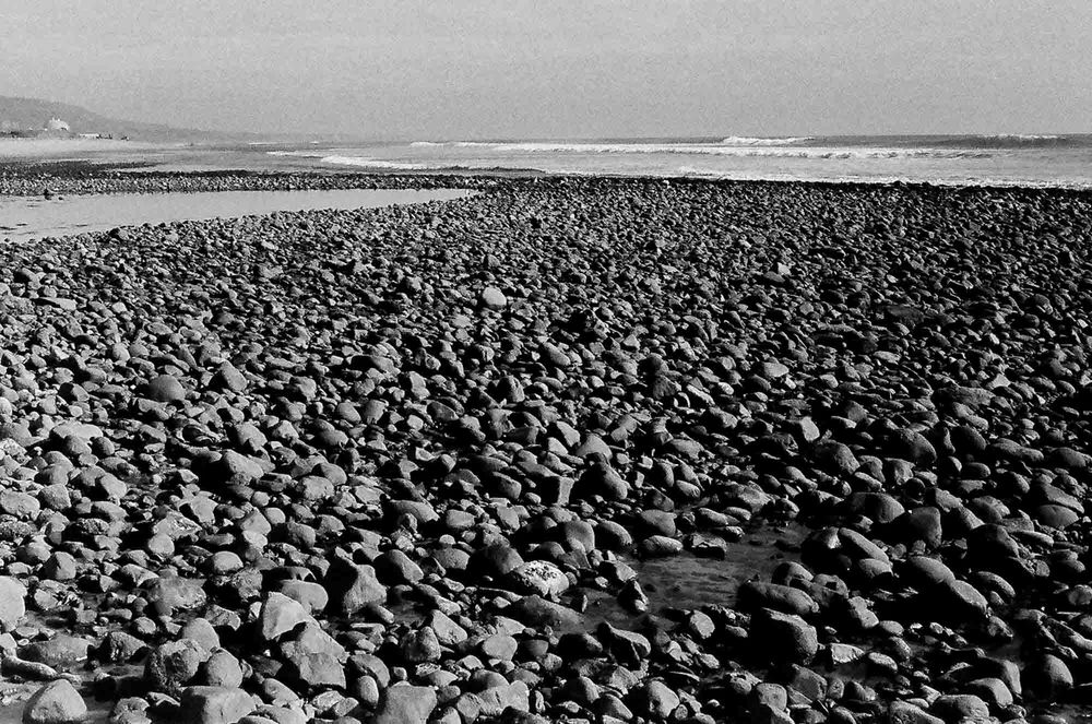 A rocky beach shore covered in countless smooth stones stretches toward the distant horizon and water.