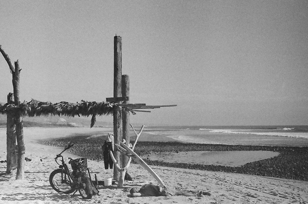 Black and white photograph of a scenic coastal beach with bicycles parked along wooden posts near the shoreline.