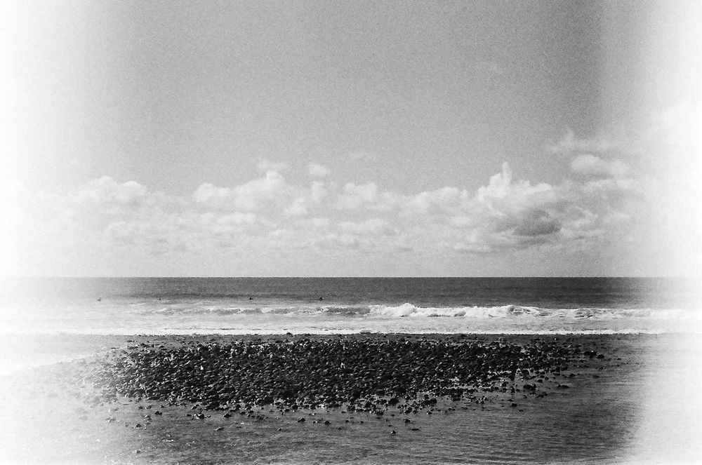 A black and white vintage photograph shows ocean waves crashing against a rocky shoreline under a cloudy sky.
