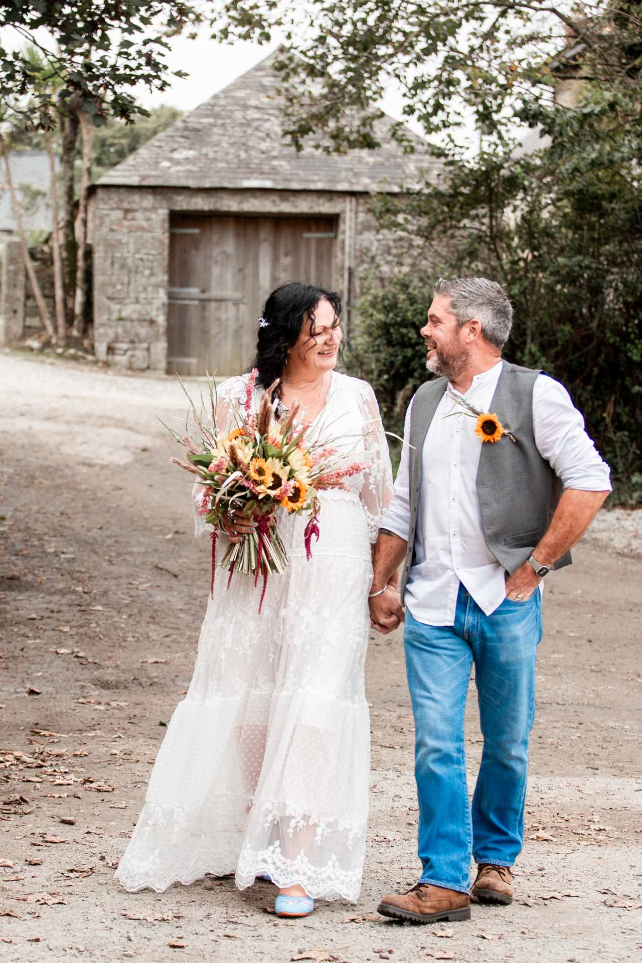 A couple in wedding attire poses outdoors with sunflowers against a rustic stone wall backdrop.