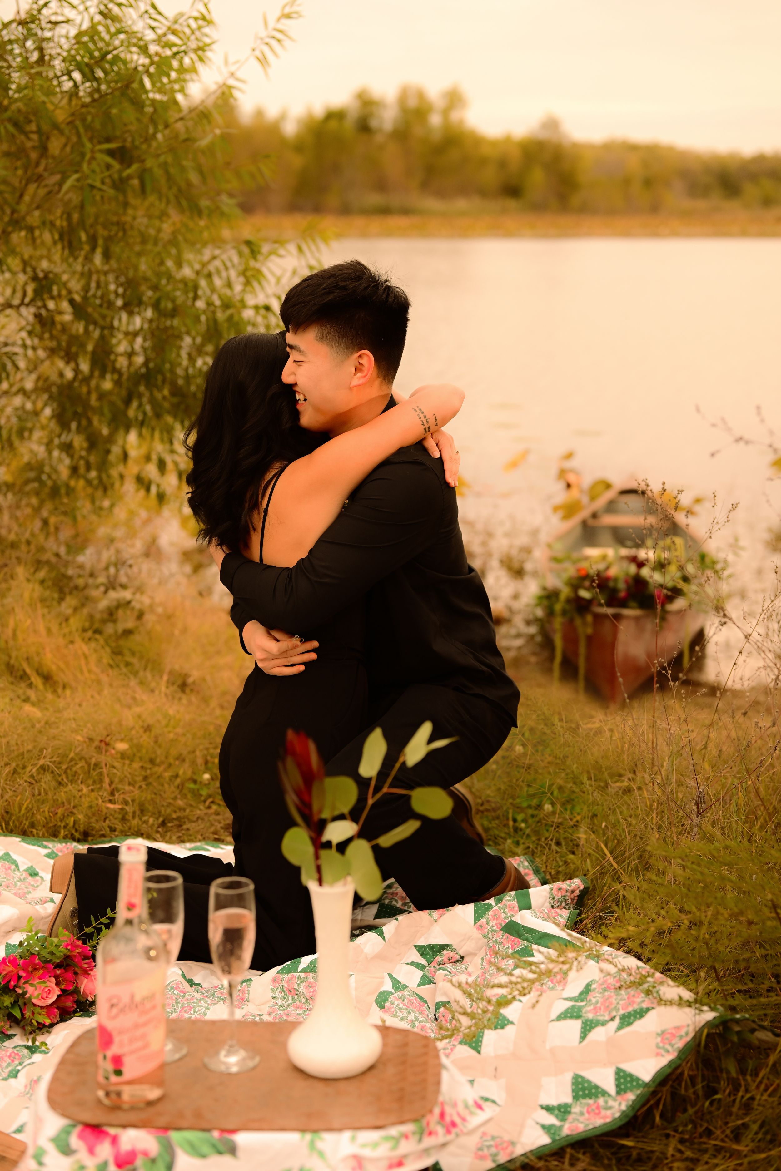 Romantic lakeside picnic setup with flowers and candles at sunset as couple embraces in the background.