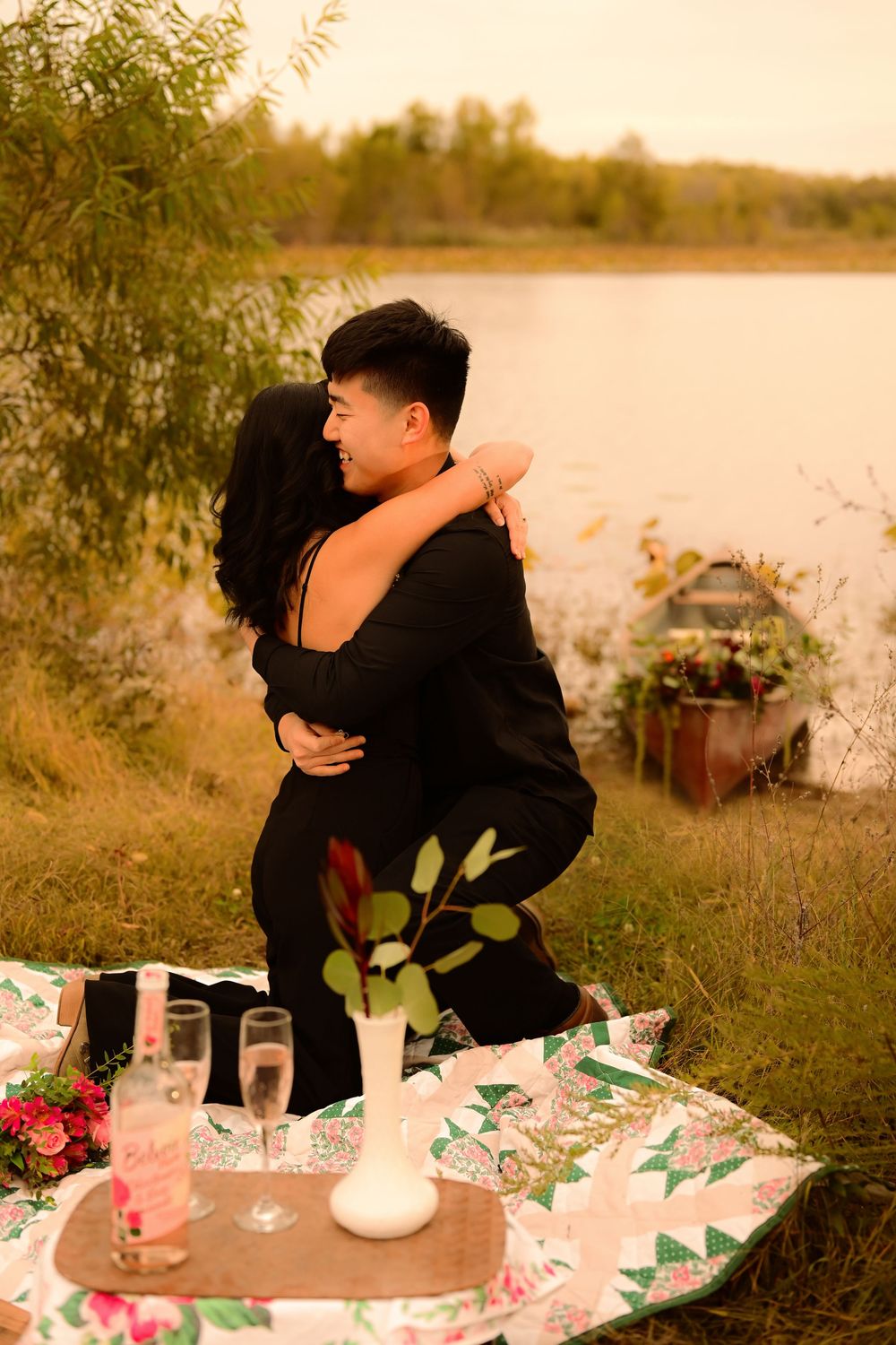 Romantic lakeside picnic setup with flowers and candles at sunset as couple embraces in the background.