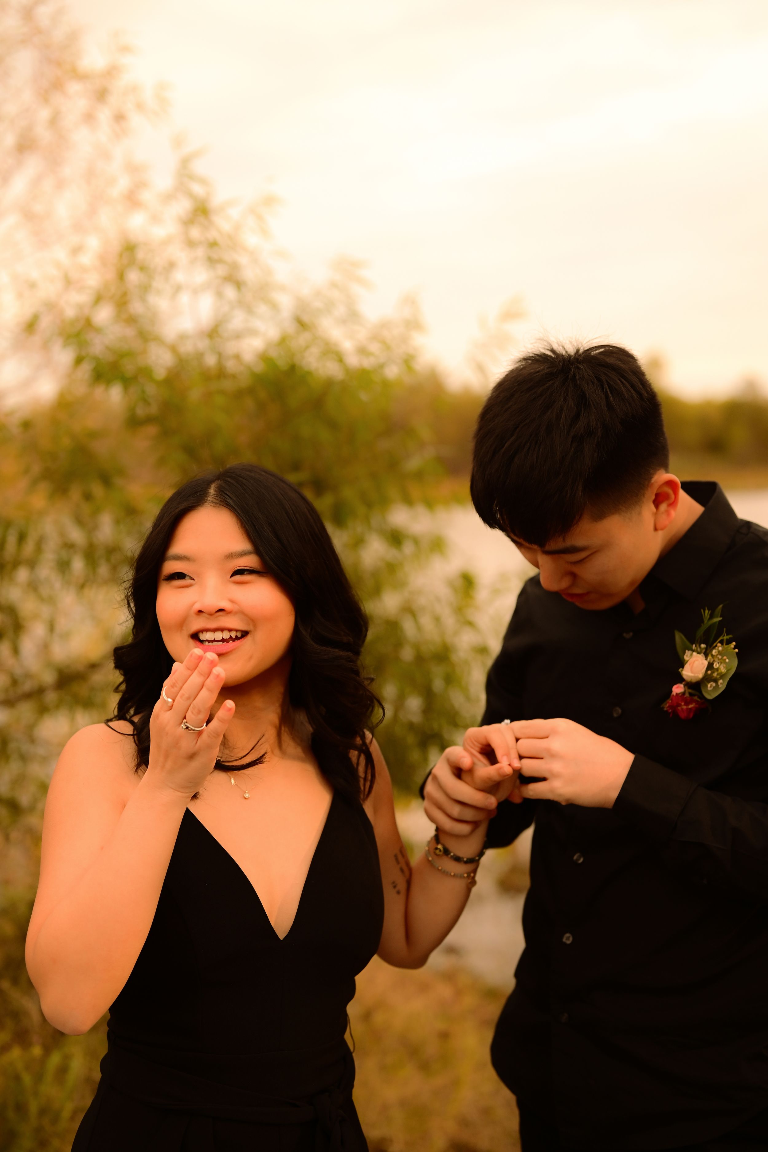 A couple shares a joyful moment in formal attire during sunset, with natural foliage in the background.