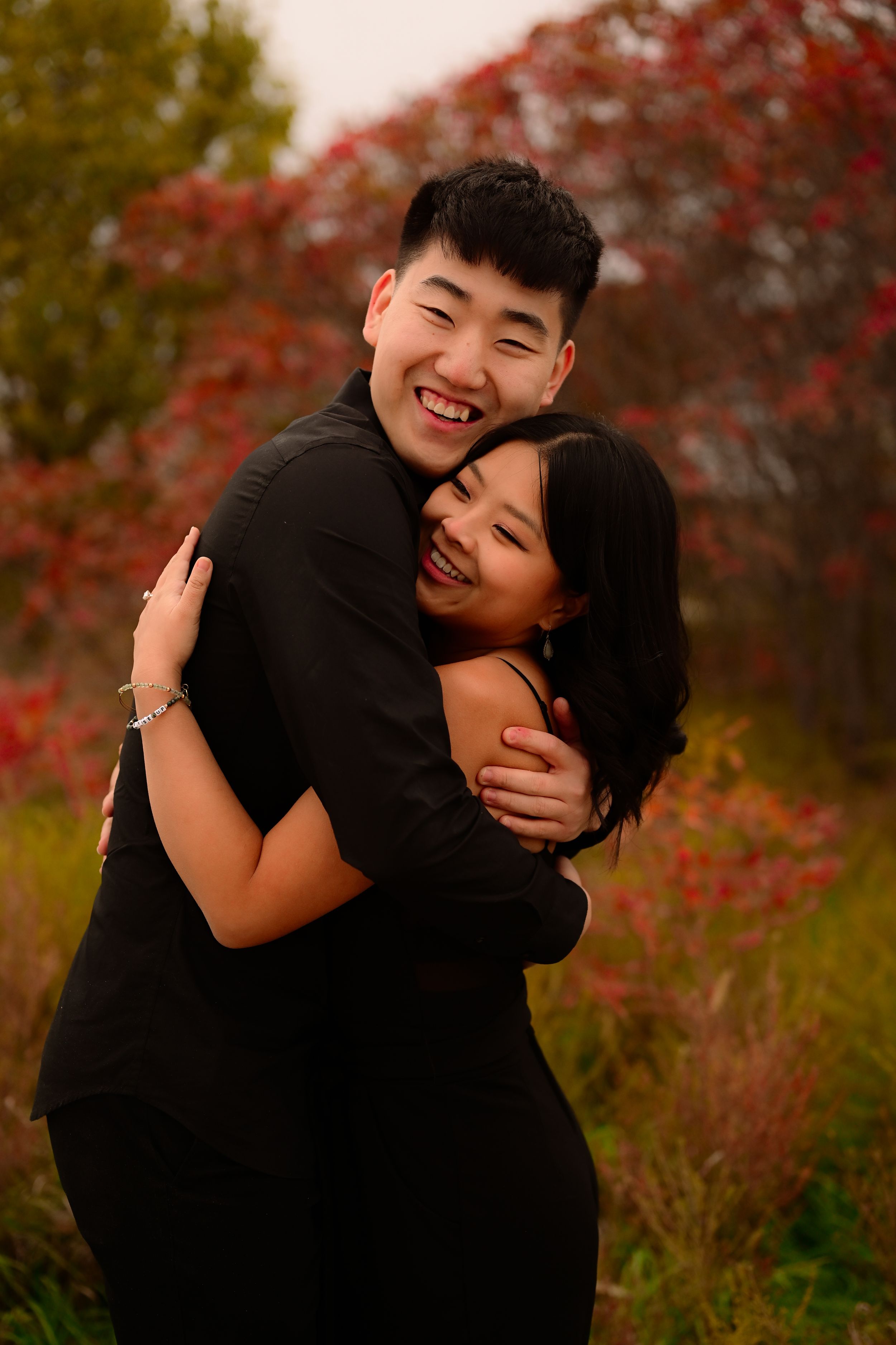 A couple embraces joyfully in a romantic autumn setting with red foliage in the background.