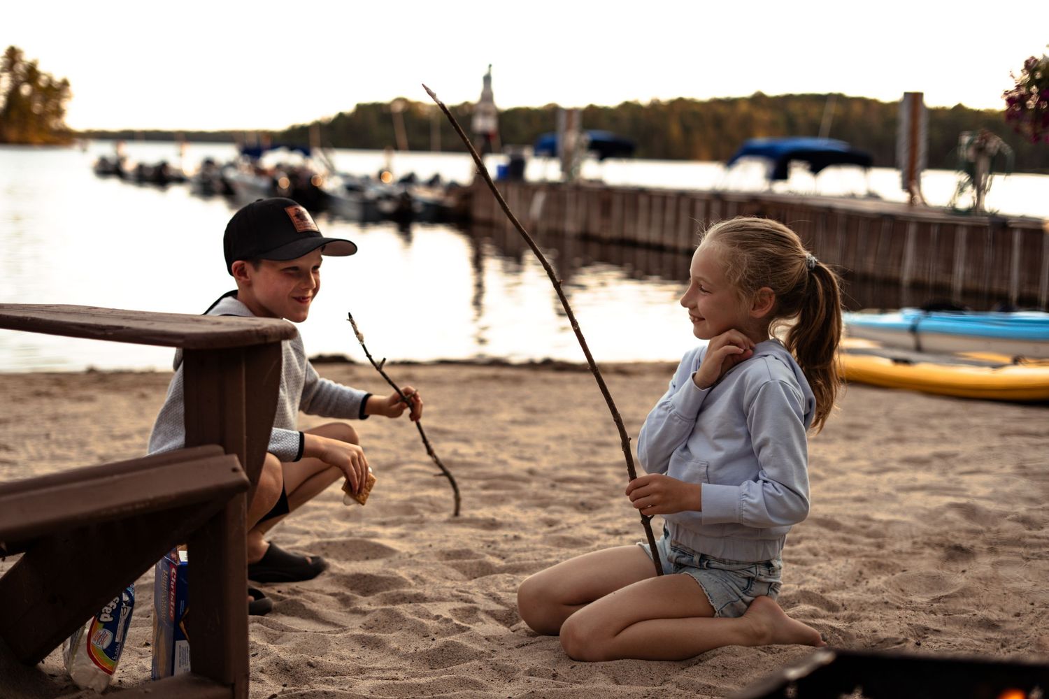 Children sitting on wooden bench swings at sunset on sandy beach.