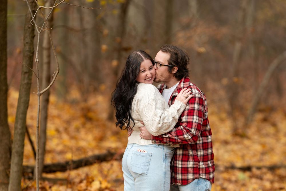 Couple sharing a kiss among golden fallen leaves in autumn forest engagement photo session.