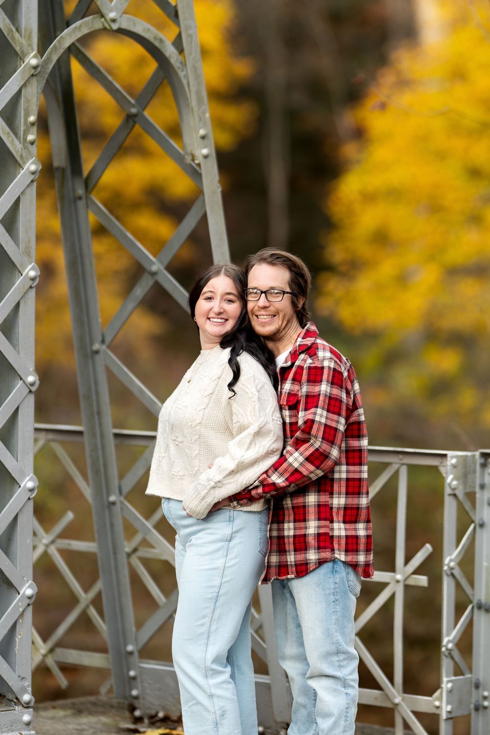 Couple embracing and smiling on a decorative metal bridge with golden autumn foliage.