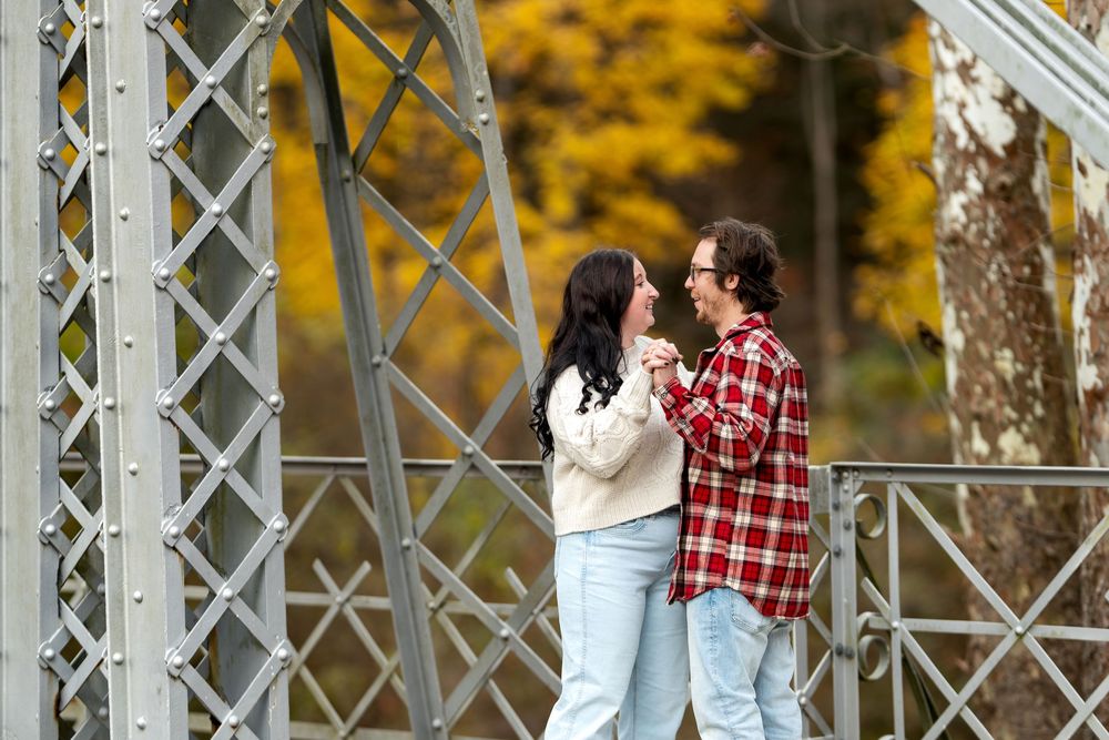 Couple holding hands on a metal bridge surrounded by colorful autumn foliage.