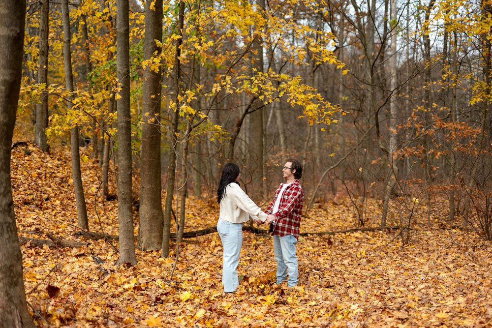 Couple holding hands in autumn forest with golden fallen leaves surrounding tall trees.