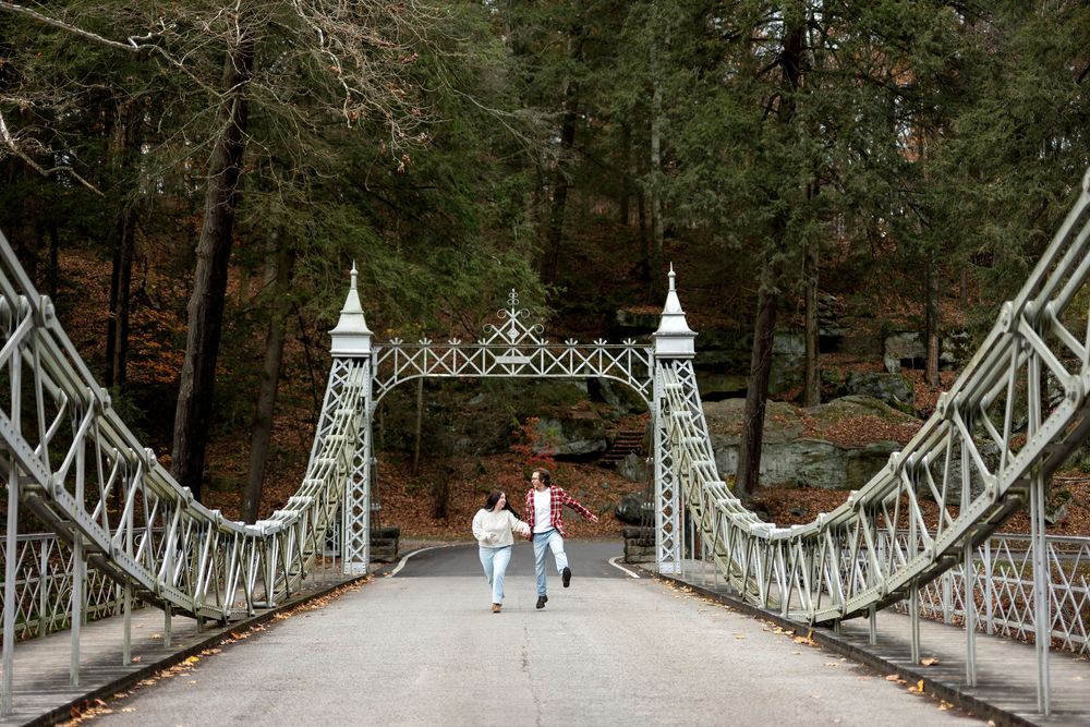 Couple walks hand-in-hand across an ornate white suspension bridge surrounded by autumn trees.
