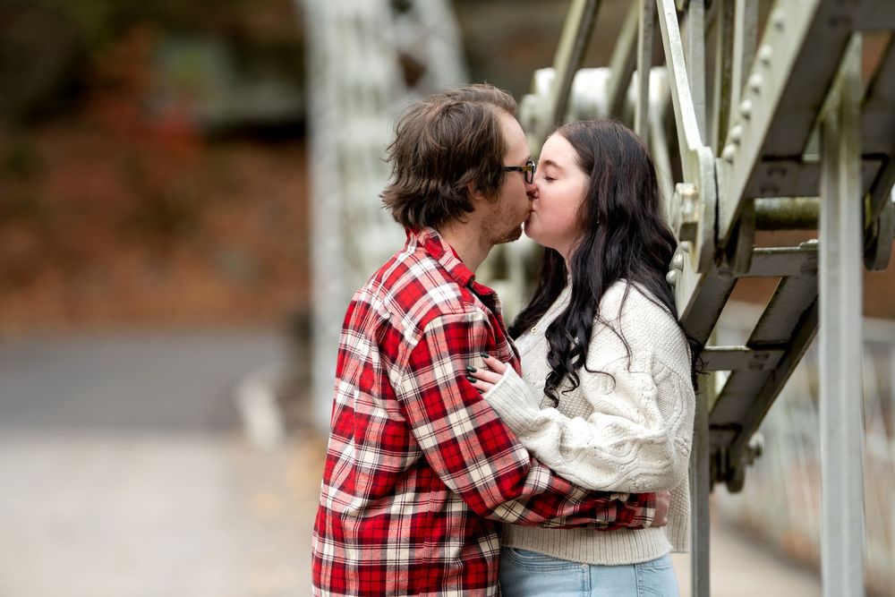 Romantic couple shares a tender kiss beside a metal bridge railing with autumn backdrop.
