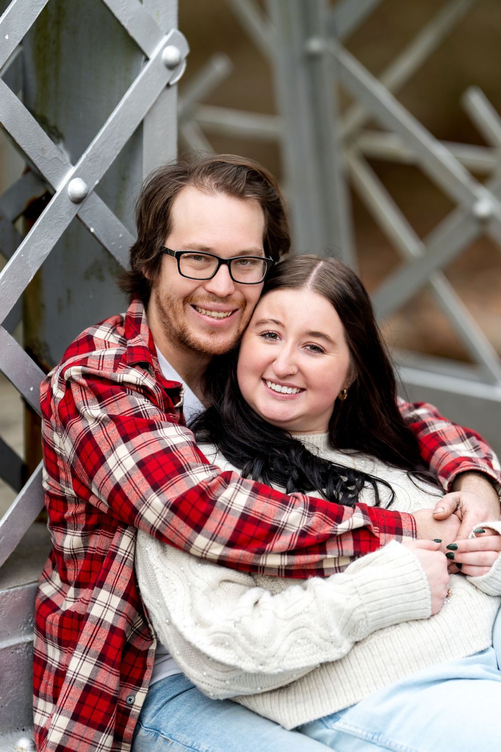 Smiling couple embraces while sitting against a metal bridge structure in a close portrait.