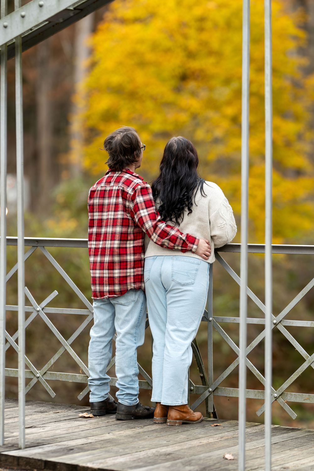 Couple stands arm-in-arm on a metal bridge overlooking vibrant yellow autumn foliage.