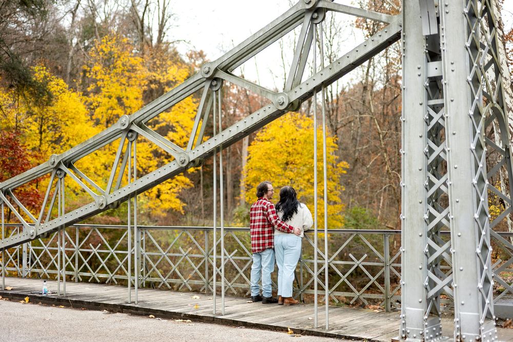 Couple stands close together on a metal truss bridge surrounded by colorful autumn foliage.