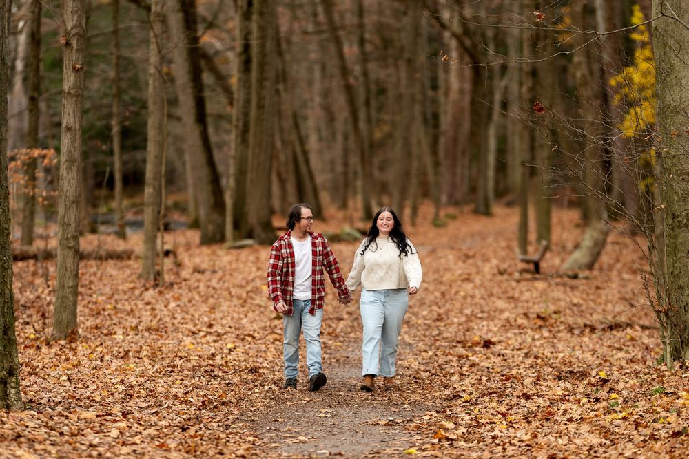 Smiling couple walking hand in hand along a forest trail covered in fall leaves.