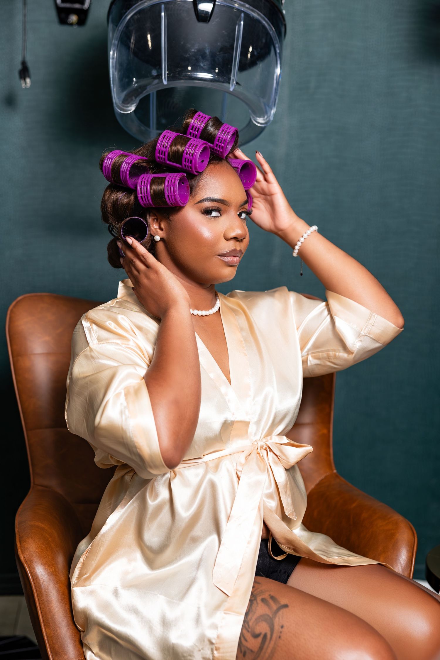 Salon scene with pink hair rollers and cream silk robe in a leather chair under a hair dryer.