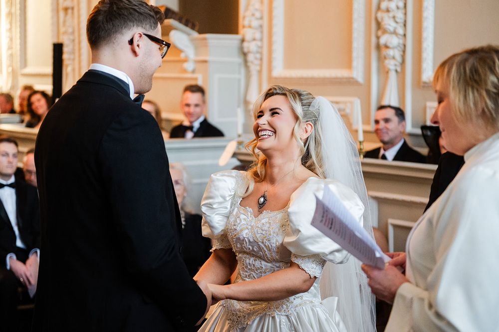 A couple shares a candid laughing moment during their wedding ceremony in a grand interior setting.
