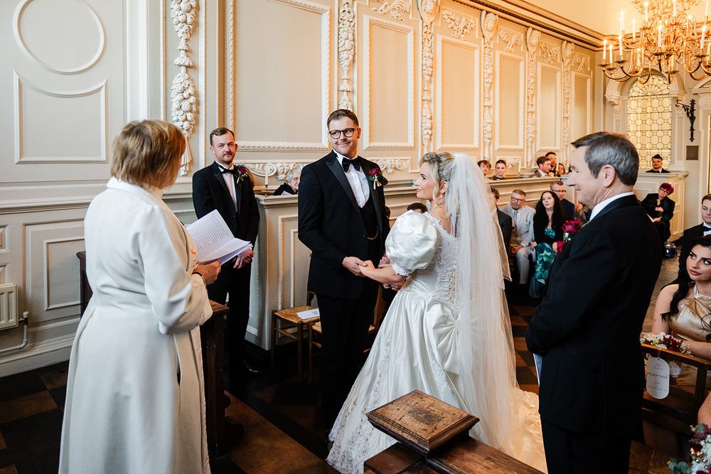 Wedding ceremony guests gather in an elegant ballroom with ornate white walls and crystal chandeliers.