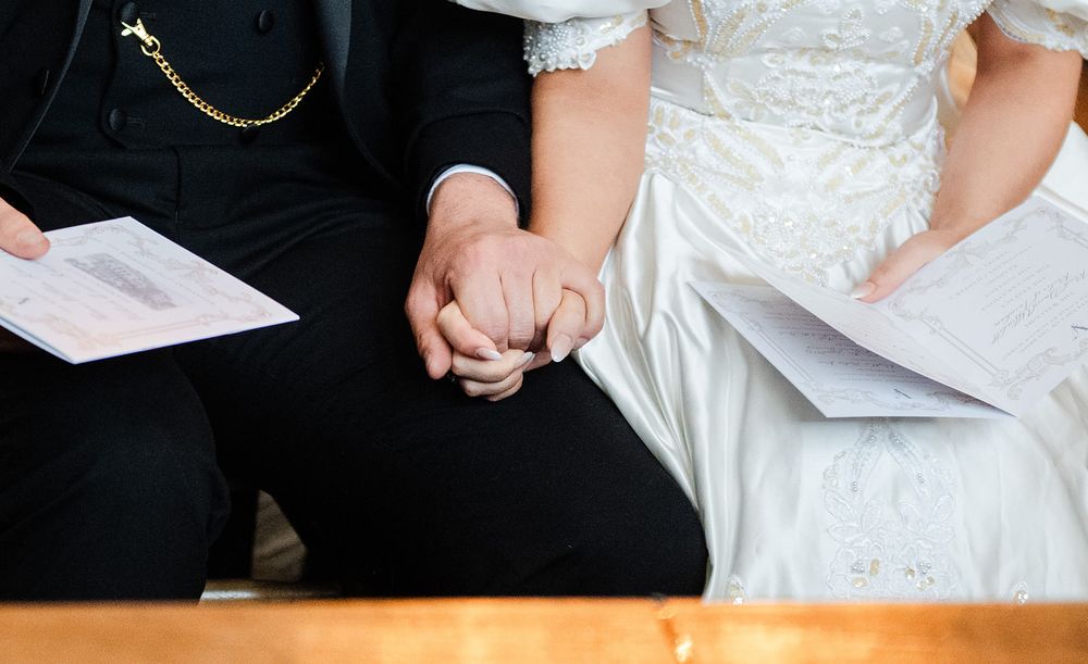 A couple signing documents at a wooden table, with hands visible on paperwork and a white dress in view.