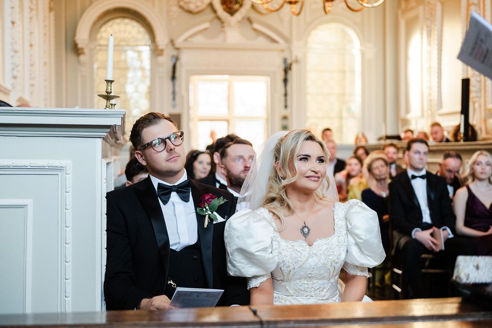 Wedding guests seated in rows at an elegant white-walled venue with arched windows and chandeliers.
