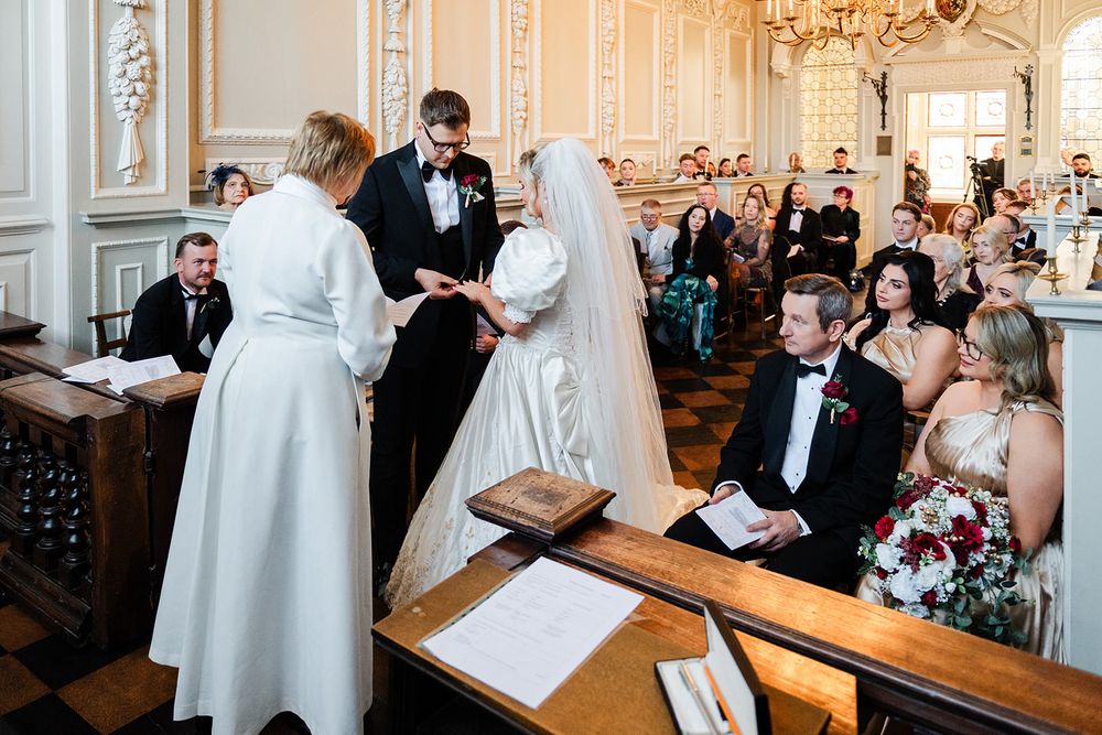 A traditional wedding ceremony takes place in an ornate chapel with white walls and chandeliers as guests look on.