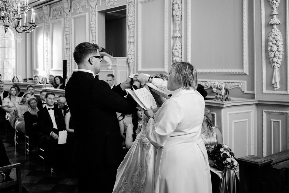 A black and white photograph of a couple exchanging rings during a traditional wedding ceremony in an ornate room.
