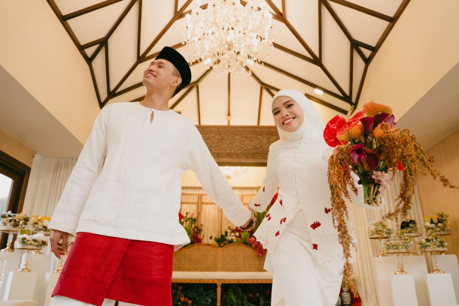 Malay couple in traditional wedding attire holding hands and laughing inside a warmly lit venue.