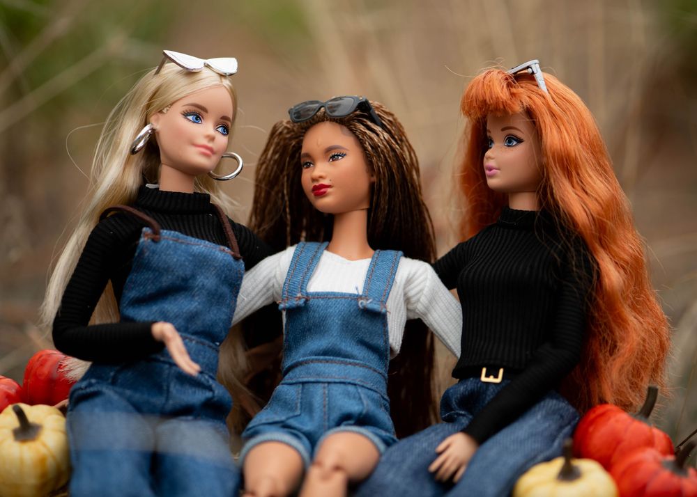 Three diverse Barbie dolls in denim outfits sitting among autumn pumpkins outdoors.