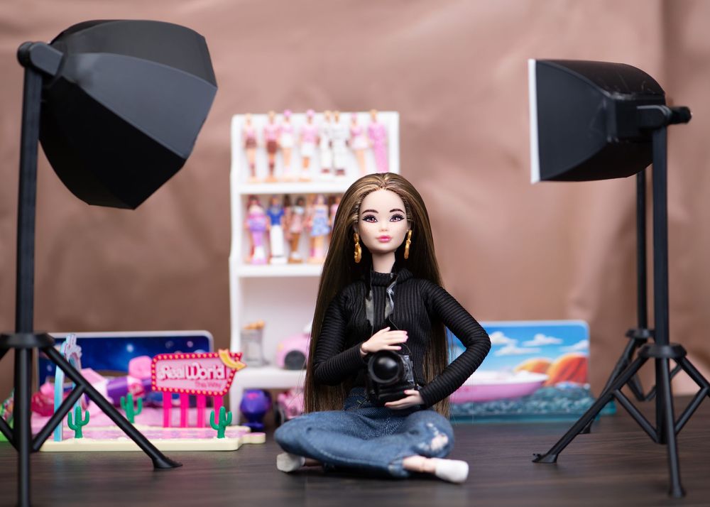 Barbie photographer doll sitting cross-legged with camera in front of a colorful toy display shelf.