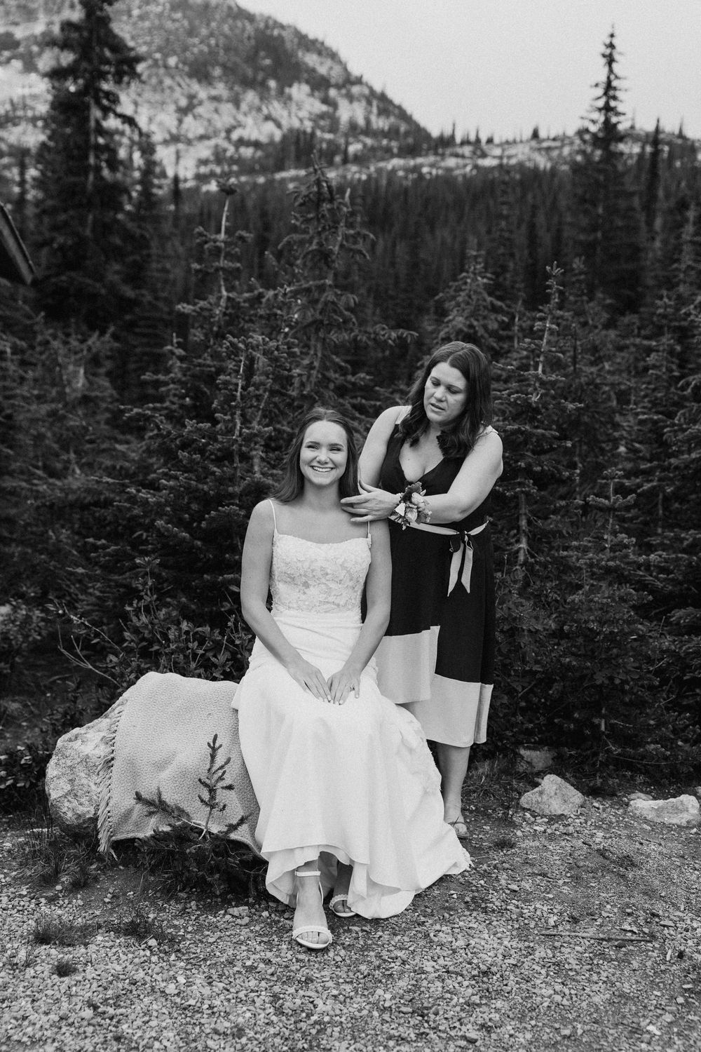 A black and white mountain wedding photo showing two people posing among evergreen trees in a scenic alpine setting.
