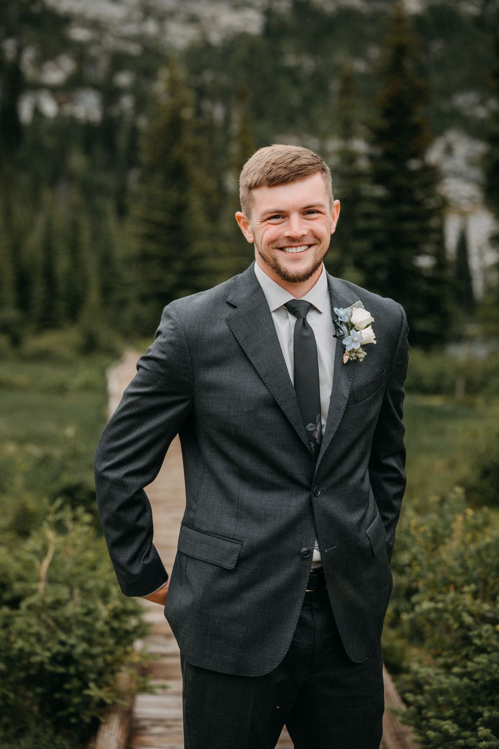 A groom in a dark suit and boutonniere smiles during an outdoor mountain wedding photoshoot.