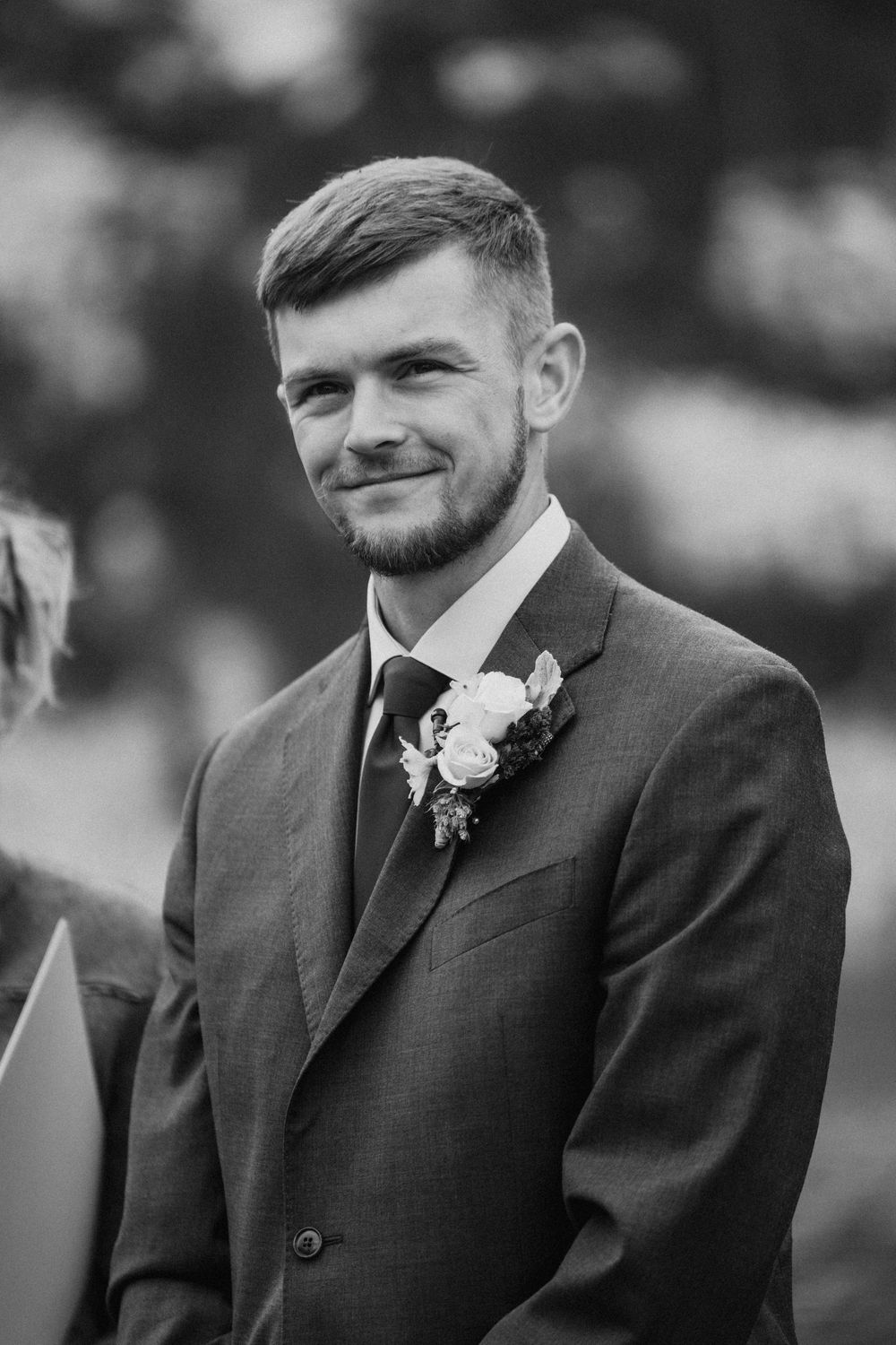A groom wearing a dark suit and boutonniere smiles in this black and white portrait.